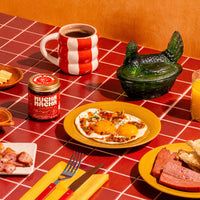 A styled breakfast table scene with a red-and-white striped ceramic mug filled with coffee, a jar of MUCHA MACHA, a plate of fried eggs, sliced sausage, toast, and orange juice on a red checkered tablecloth.