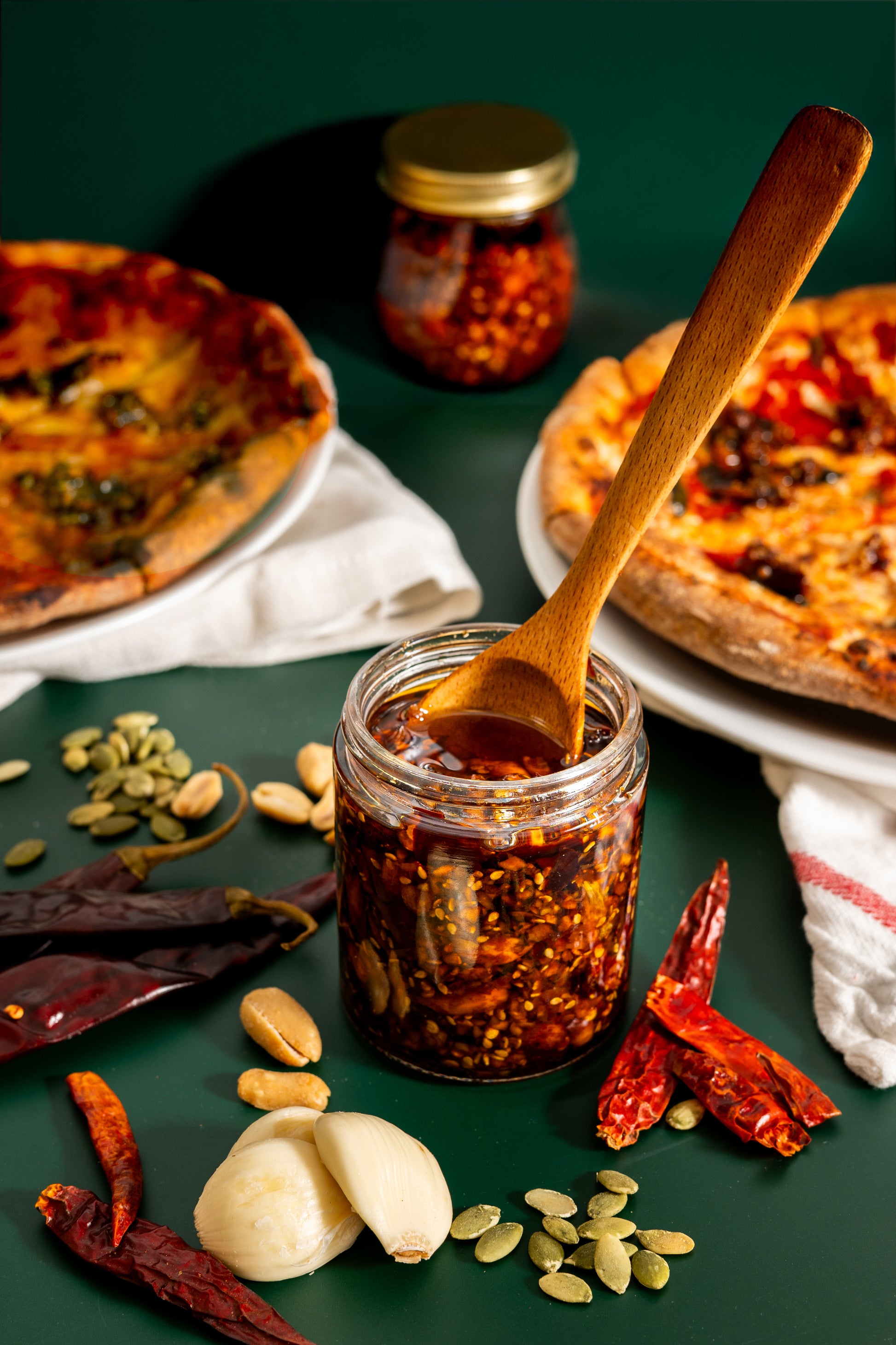 Jar of red pepper flakes with a wooden spoon, surrounded by pizzas and ingredients on a green surface.