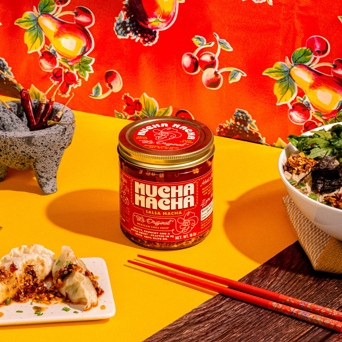 A jar of Mucha Macha is displayed on a bright yellow table, surrounded by food props including dumplings, a bowl of noodles, chopsticks, and a stone mortar and pestle, with a vibrant red fruit-patterned backdrop.