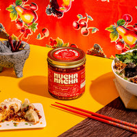A jar of Mucha Macha is displayed on a bright yellow table, surrounded by food props including dumplings, a bowl of noodles, chopsticks, and a stone mortar and pestle, with a vibrant red fruit-patterned backdrop.