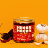 A jar of Mucha Macha Salsa Macha centered on a bright yellow-orange background, styled with dried red chilies, garlic bulbs, and loose garlic cloves around it.