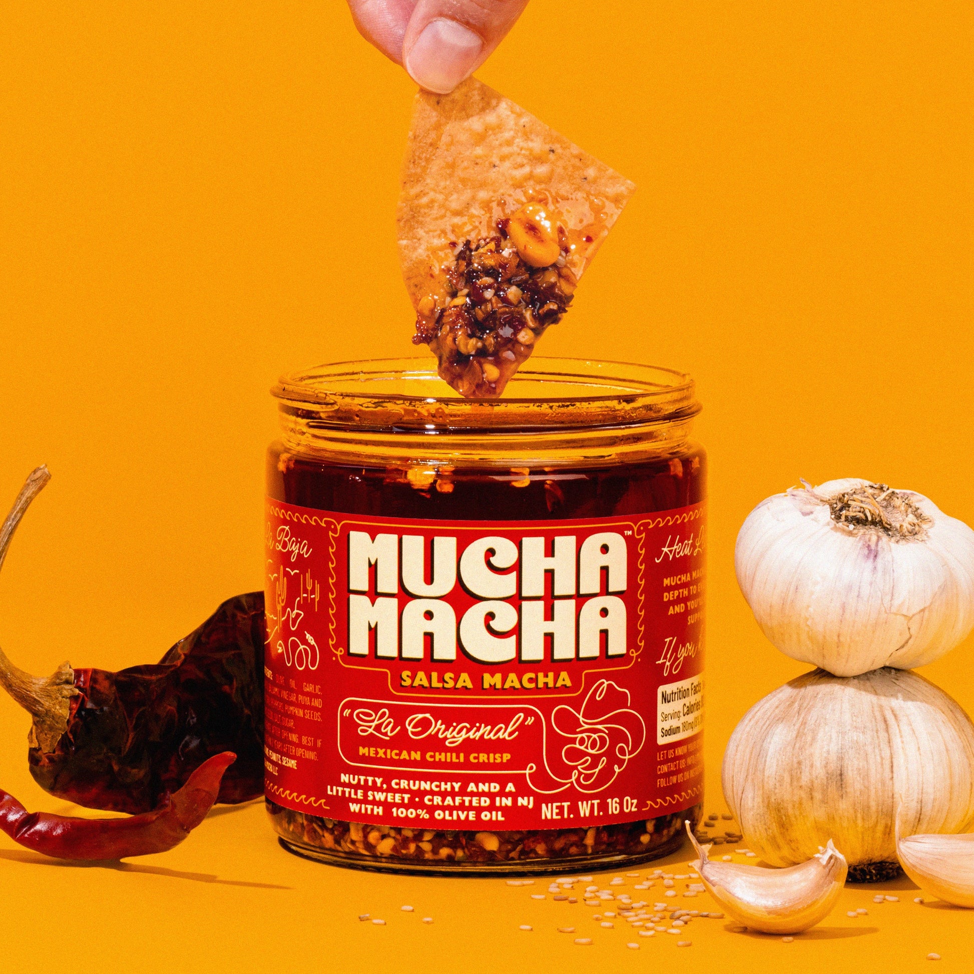 A jar of Mucha Macha Salsa Macha is shown on a bright yellow background, with a hand dipping a tortilla chip into the sauce. Whole garlic bulbs, dried red chilies, and scattered sesame seeds surround the jar.