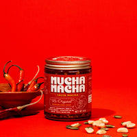 A jar of Mucha Macha Salsa Macha with olive oil and seeds, styled on a bright red background with chili peppers in a wooden bowl and scattered seeds in the foreground.
