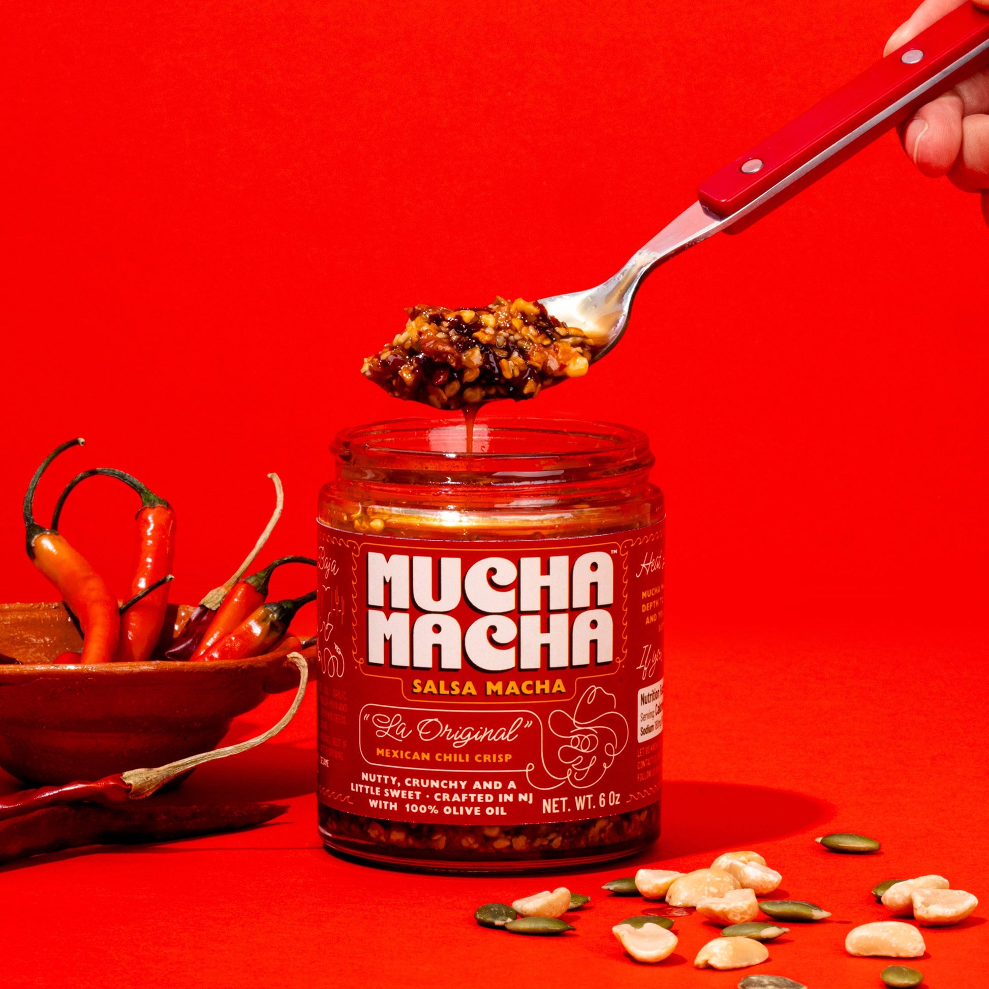 A jar of Mucha Macha on a bright red background, with a hand holding a spoonful of the sauce above the jar. A bowl of red chilies and scattered peanuts are visible on either side.