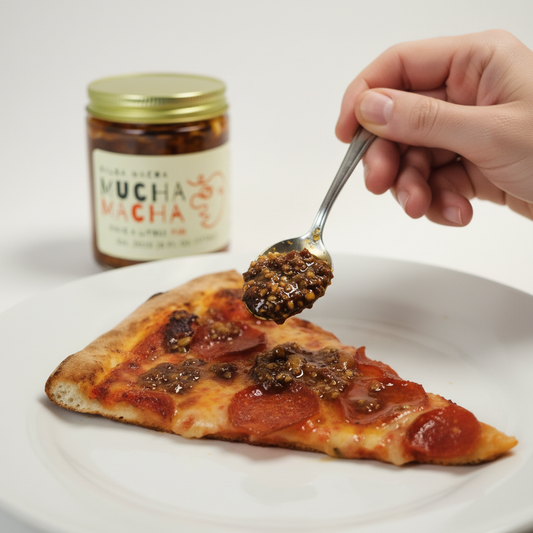 Spoon adding Mucha Macha to a slice of pizza with a jar labeled 'Mucha Mucha' in the background.