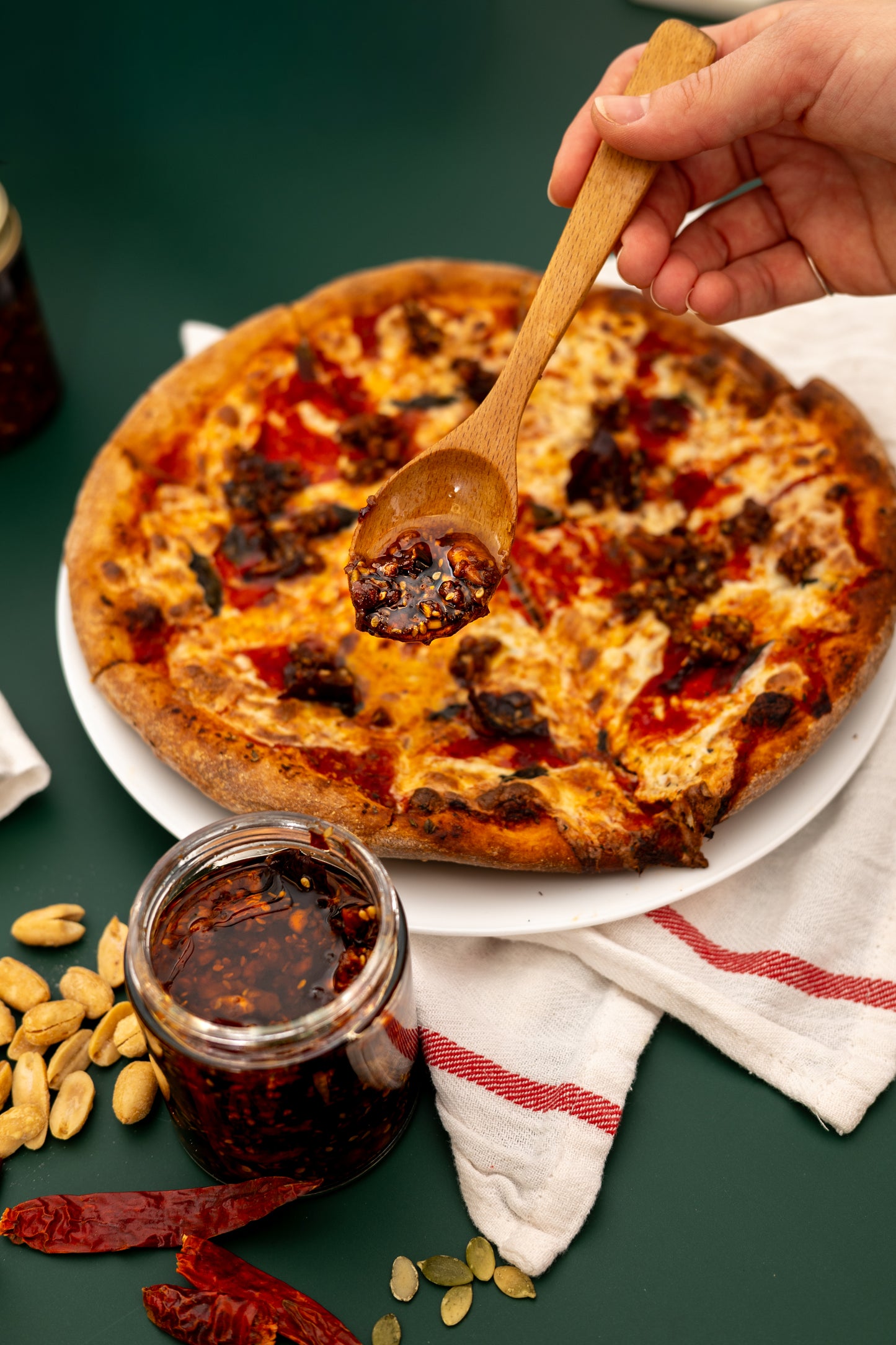Wooden spoon scooping sauce from a jar over a pizza on a green surface with ingredients around.