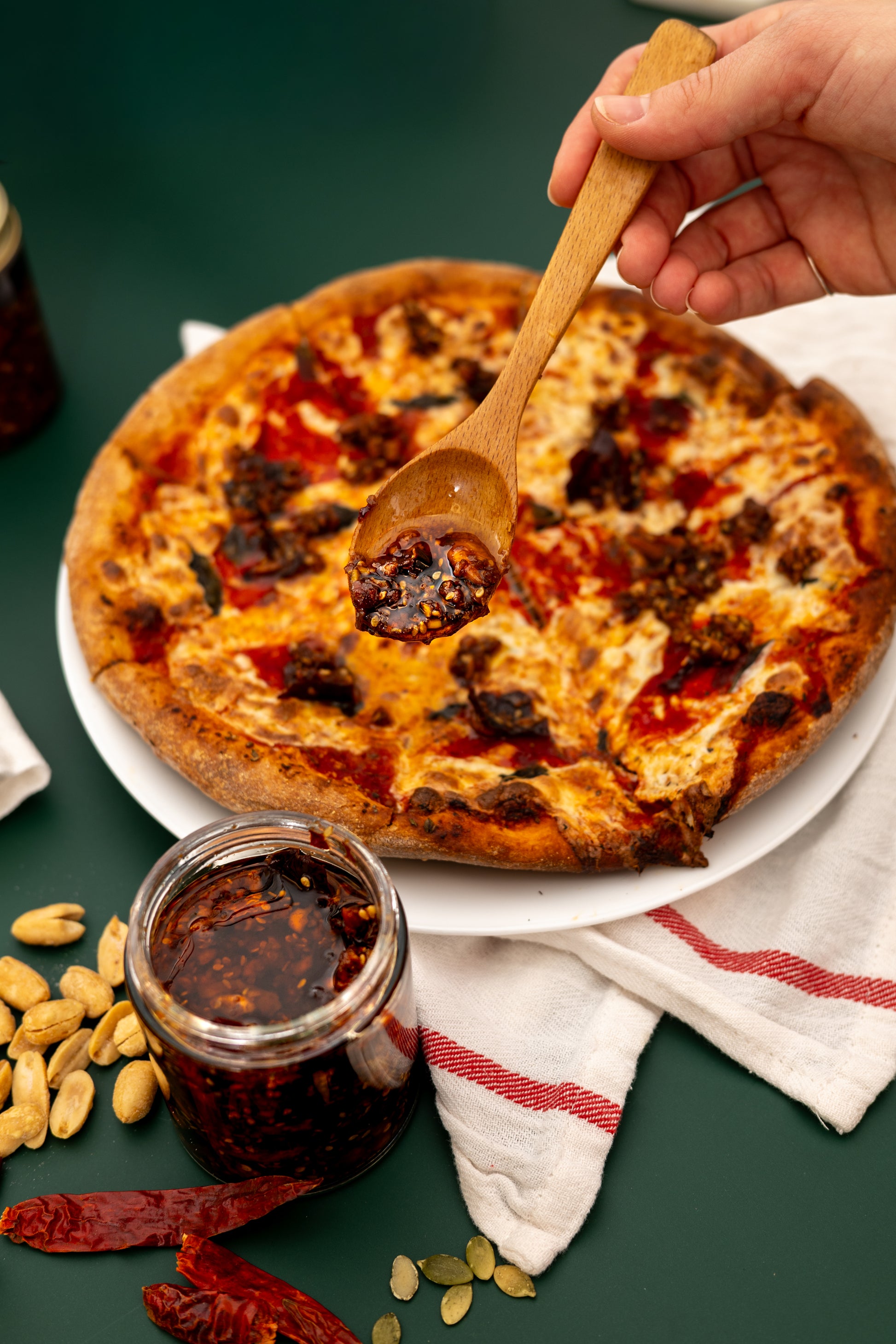 Wooden spoon scooping sauce from a jar over a pizza on a green surface with ingredients around.