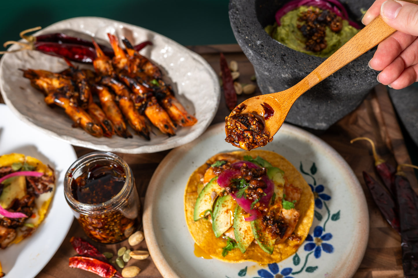 Plates of food including grilled shrimp, a tostada, and a jar of sauce on a wooden table.