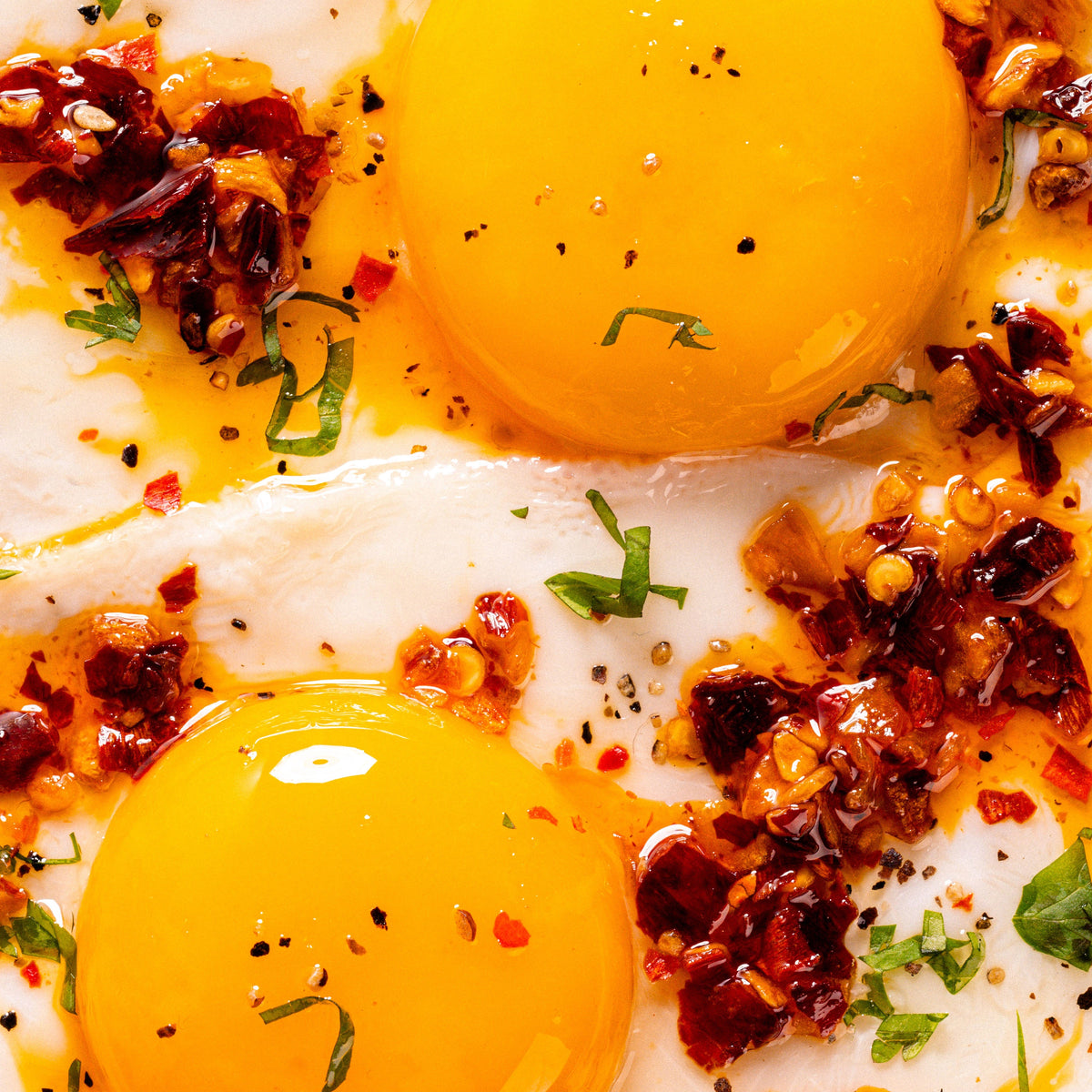 Close-up of two sunny-side-up eggs topped with MUCHA MACHA, and green herbs on a white plate.