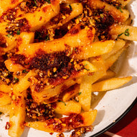 Close-up of a plate of golden french fries topped with Mucha Macha, crushed garlic, and sesame seeds.