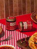 Two jars of Mucha Macha Salsa Macha displayed on a red and white checkered tablecloth next to a large Caesar salad and pizza slices.